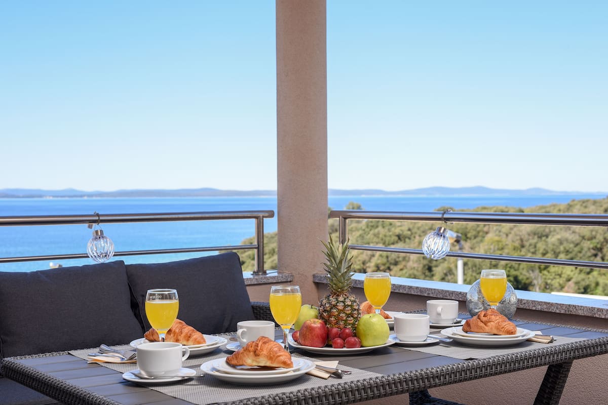 A breakfast table with croissants, fruit, and juice on a balcony overlooking a scenic view.