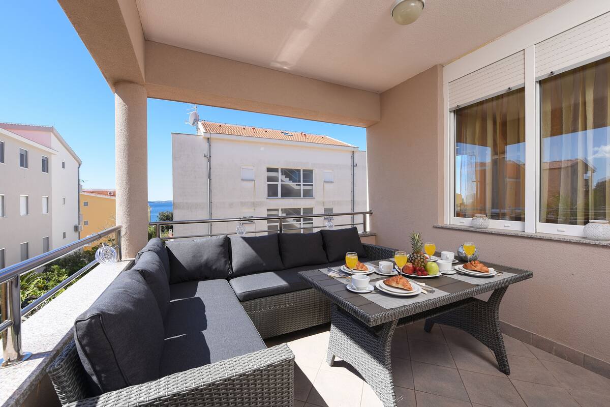 A balcony with a wicker sofa and table, featuring breakfast items and a view of buildings.