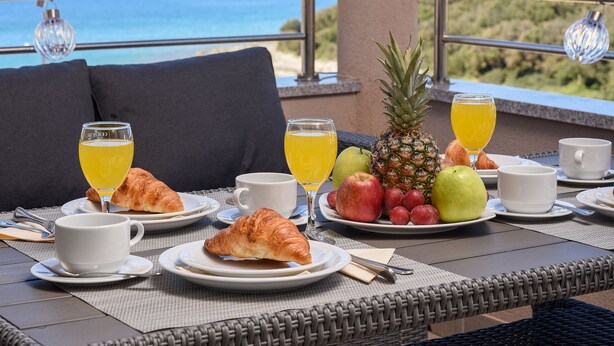A breakfast table with croissants, fruit, and juice, overlooking a scenic view. - Sunset Beach Suites (Zadar)