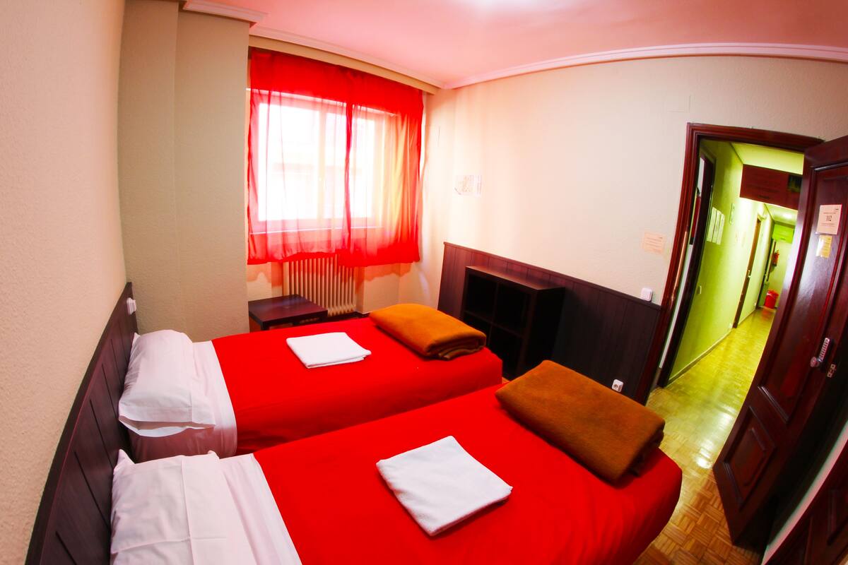 A hotel room with two beds, red bedding, a wooden headboard, a window with red curtains, and a wooden floor.