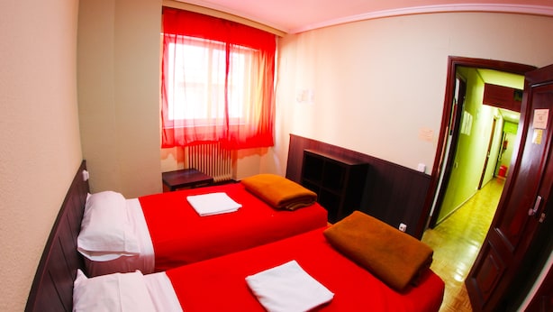 Habitación de hotel con dos camas, ropa de cama roja, cabecero de madera, una ventana con cortinas rojas y suelo de madera.