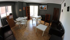 Una habitación con una mesa blanca, sillas, un sofá negro, una estantería de madera y un televisor en la pared.