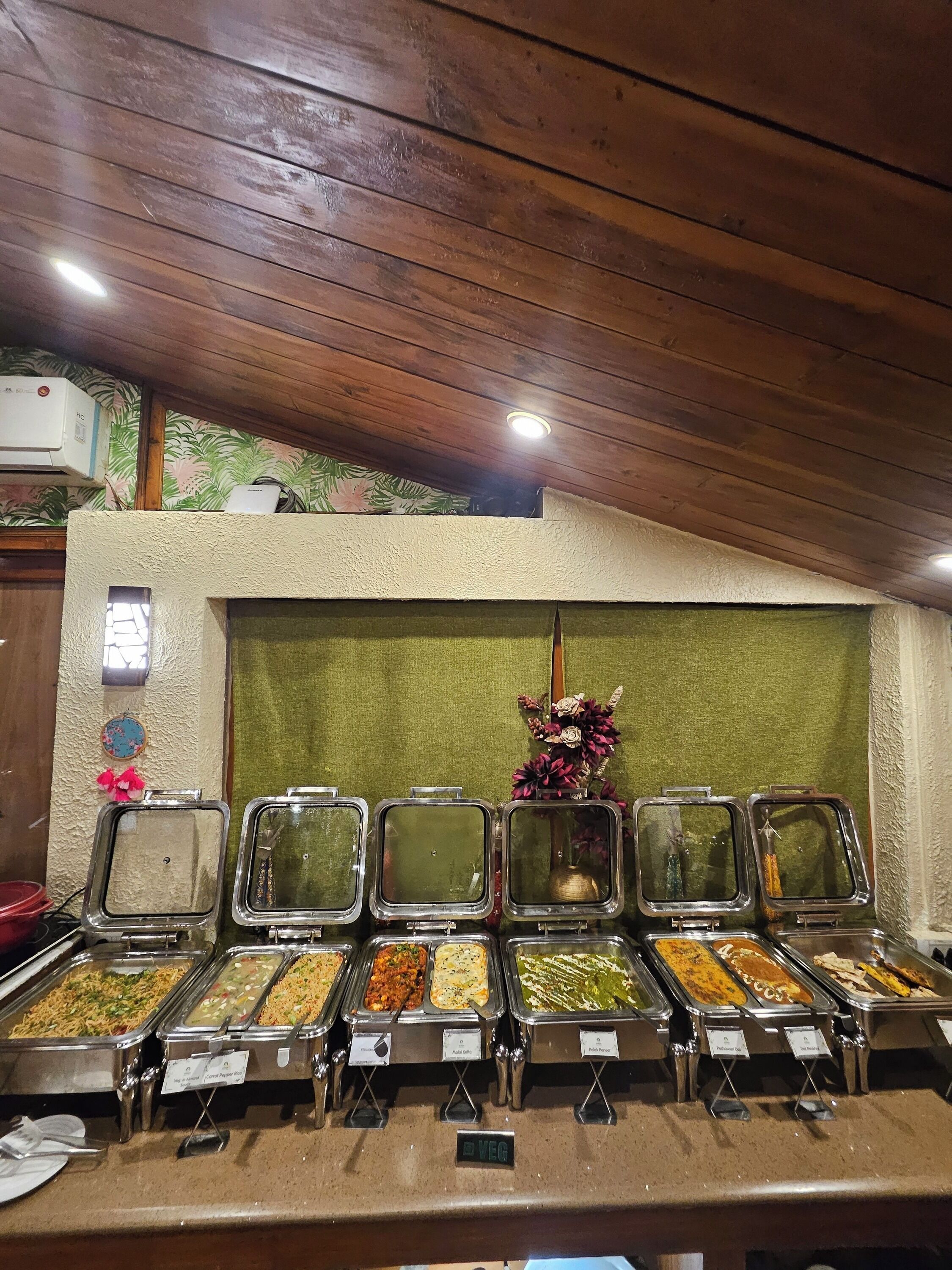 Un bufé con diversas opciones de comida, incluyendo una sección vegetariana, bajo un techo de madera con un diseño floral en la pared.