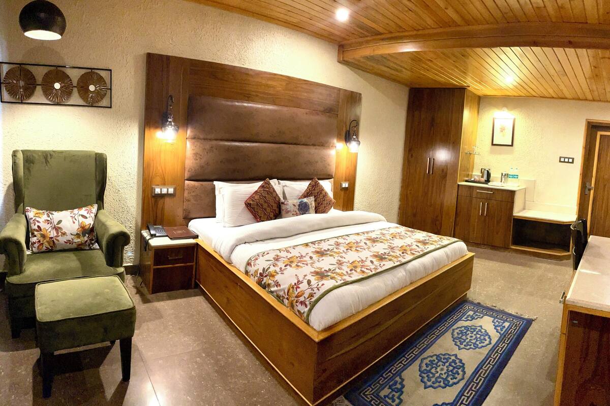 A bedroom with a wooden headboard, a green armchair, a bedside table with a lamp, a bed with floral bedding, and a small bathroom with a sink and mirror.