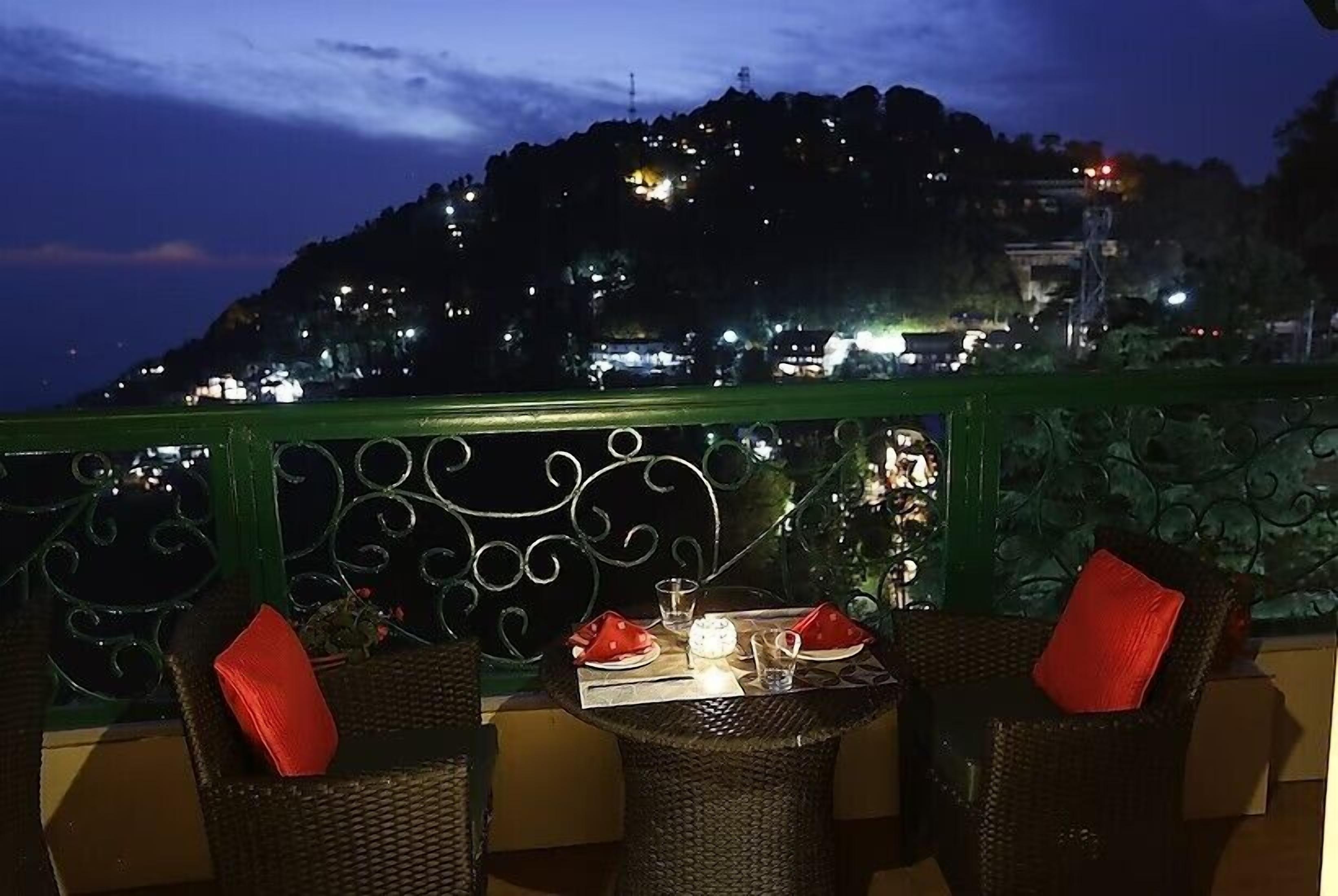 A terrace with wicker furniture and red cushions, overlooking a hillside with lights.