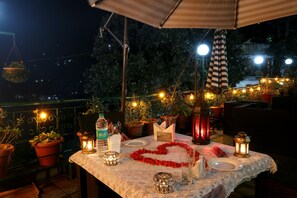 An outdoor dining area with a table set for two, surrounded by potted plants and string lights. - Hotel Mongas (Dalhousie)