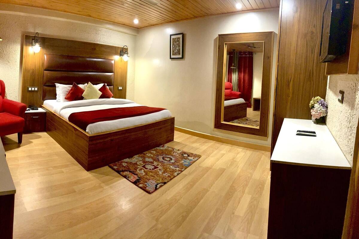 A bedroom with a wooden headboard, a bed with red and white bedding, a red armchair, a bedside table with a bottle and glasses, and a mirror on the wall.