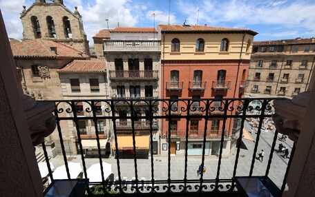 A balcony with wrought iron railings overlooking a plaza with historic buildings, including a church tower.