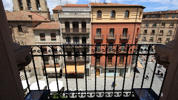 A balcony with wrought iron railings overlooking a plaza with historic buildings, including a church tower.