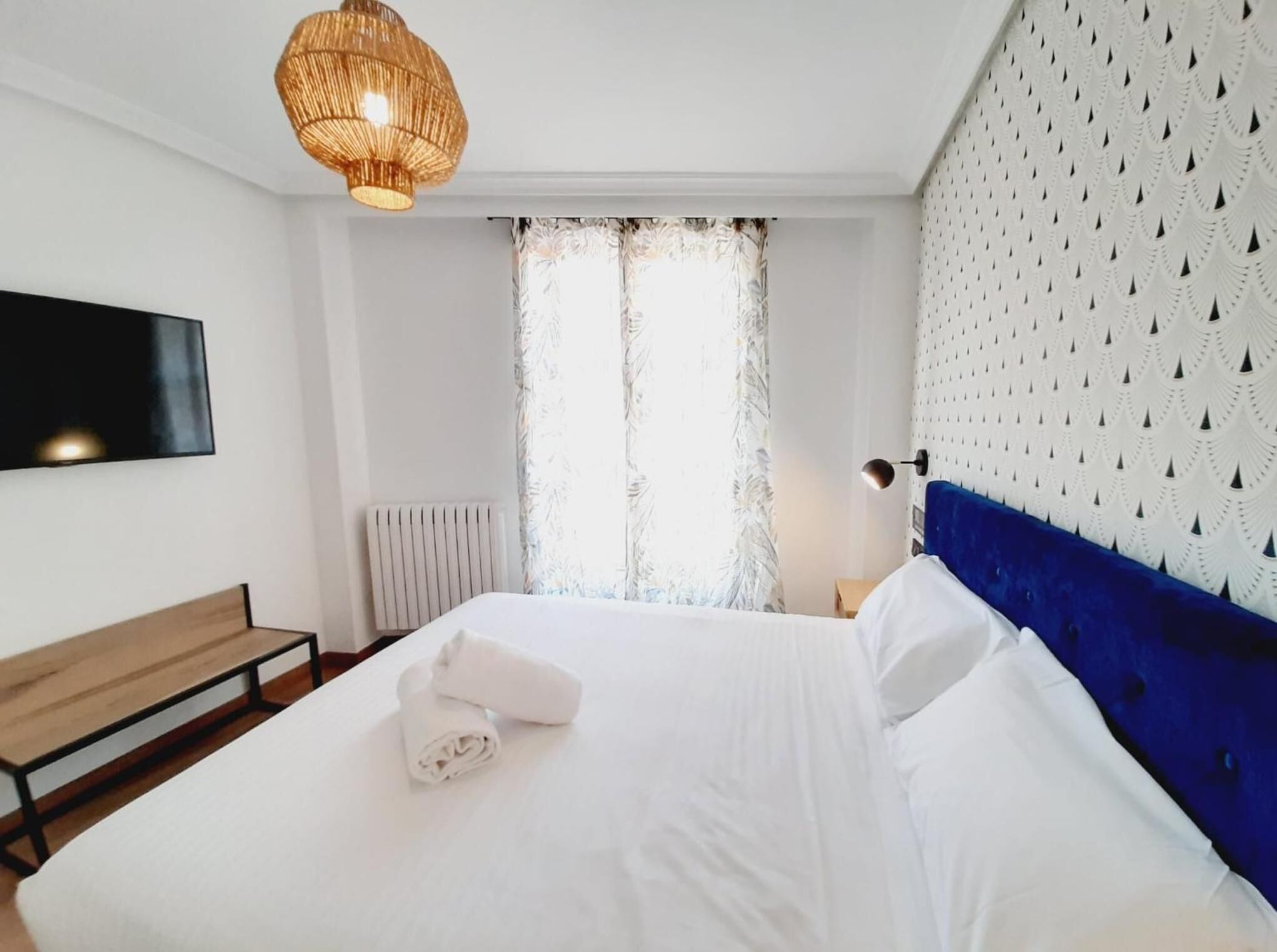Habitación de hotel con una cama grande, un banco de madera, un televisor montado en la pared, un radiador y un papel tapiz estampado.