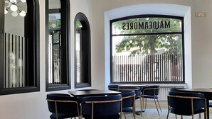 A restaurant interior with black and gold chairs, a large window with a view of a fence, and a sign reading "MADAMONDES".