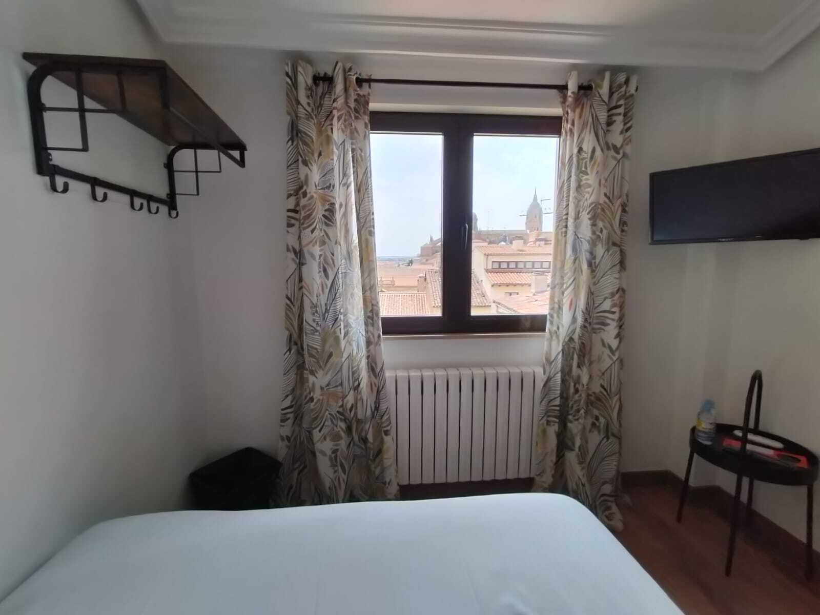 Quarto de hotel com cama, janela com cortinas, radiador, cadeira e TV fixada na parede.