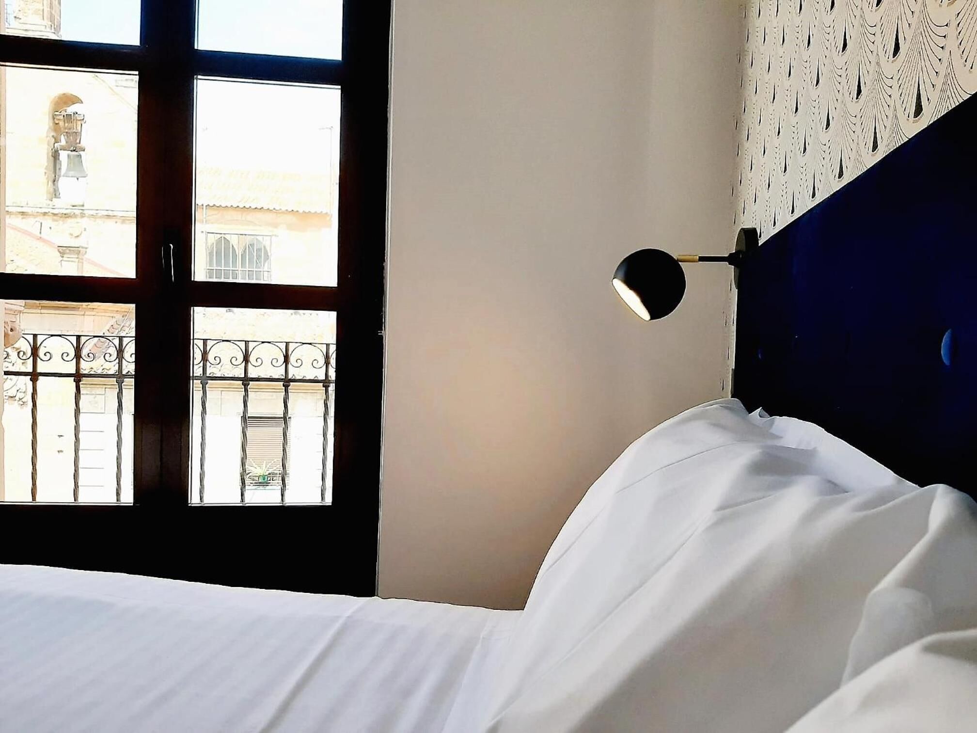 A hotel room with a bed, a window with a view of buildings, and a wall with a patterned design.