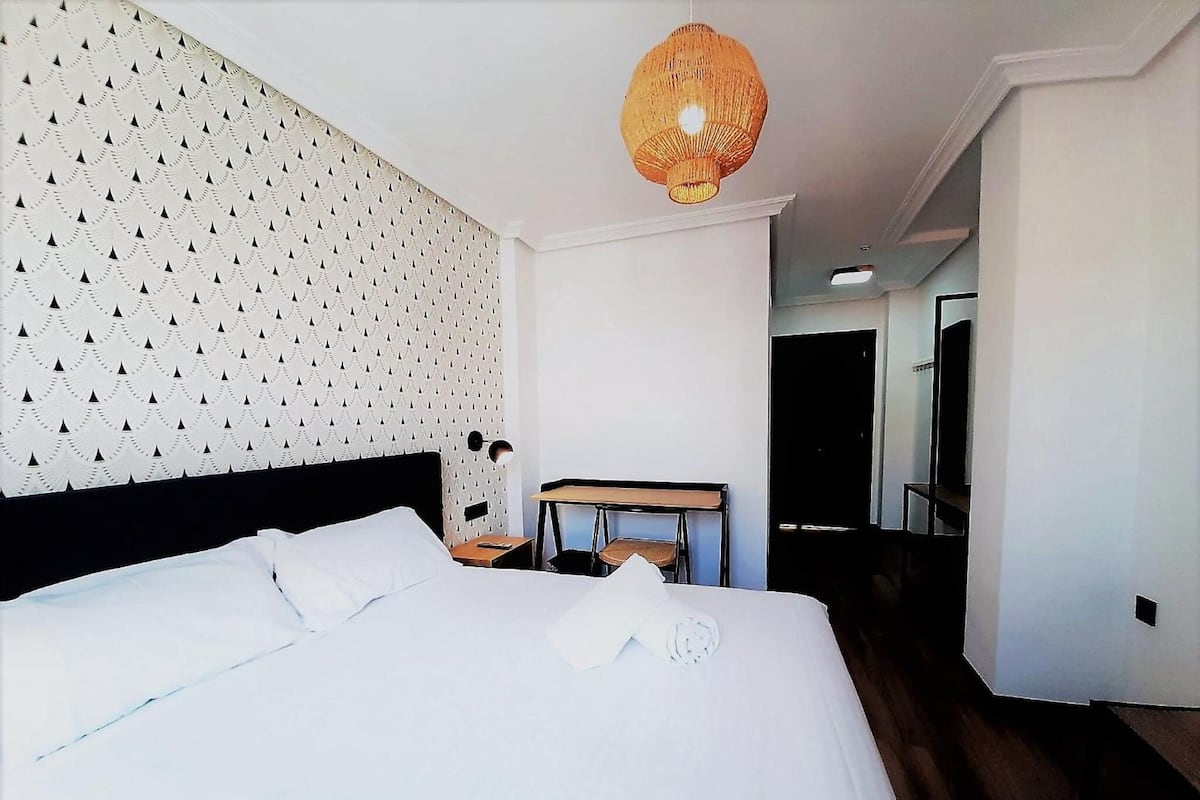 Ein Hotelzimmer mit einem ordentlich bezogenen Bett, einem kleinen Tisch mit Stuhl, einer Hängelampe und einem Spiegel an der Wand.