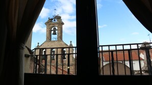 A view through a window with curtains, featuring a bell tower and a building with a red-tiled roof.