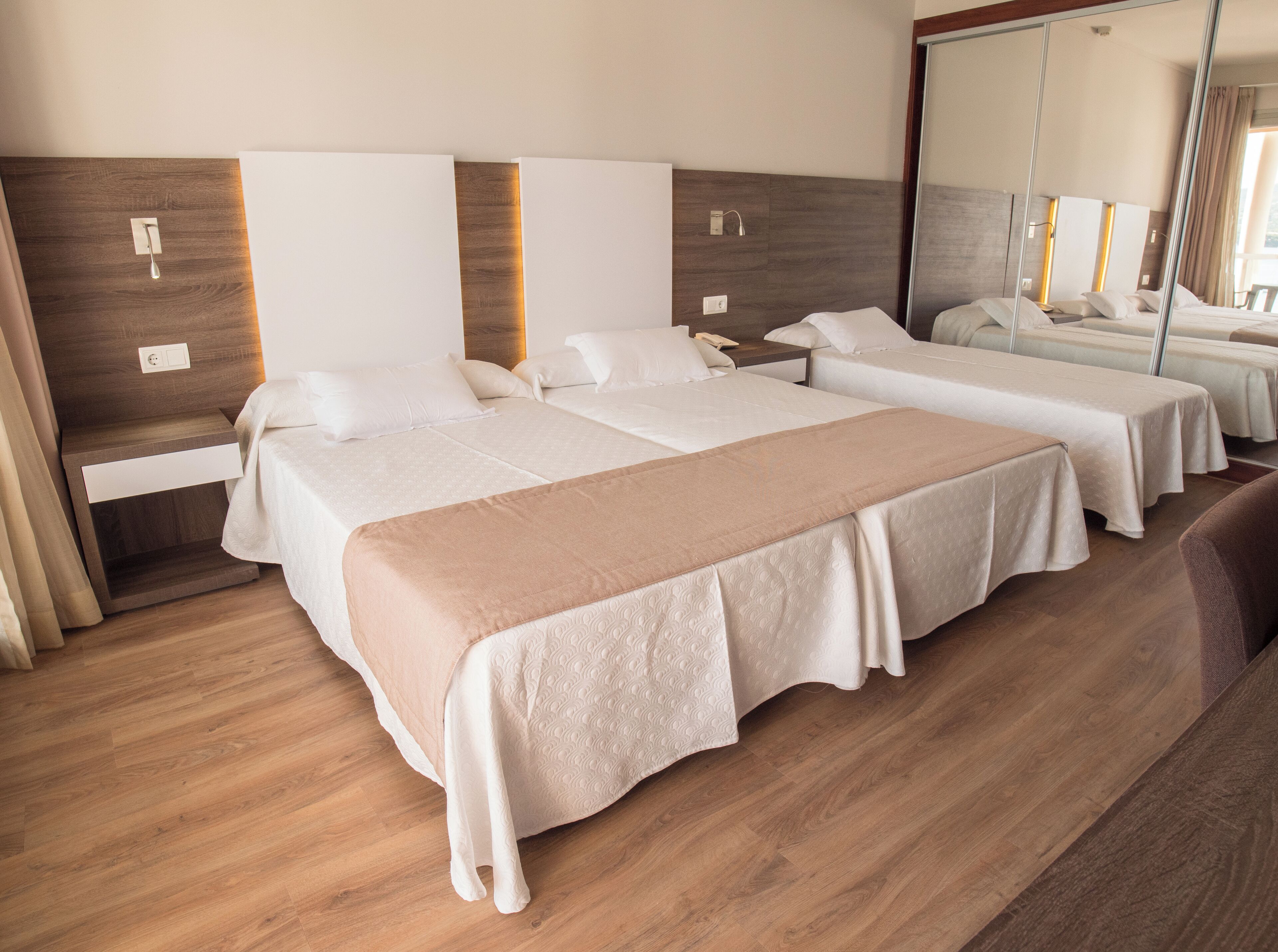 A hotel room with three beds, wooden flooring, and a large mirror on one wall.