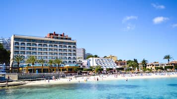 Un hotel frente al mar con varios pisos, una playa en primer plano y palmeras.