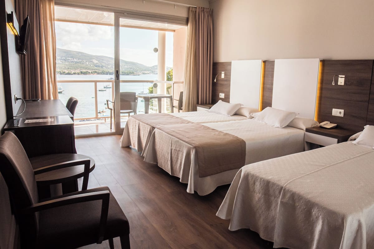 Habitación de hotel con dos camas, un escritorio y una silla. Tiene un balcón con vista al mar y a barcos.