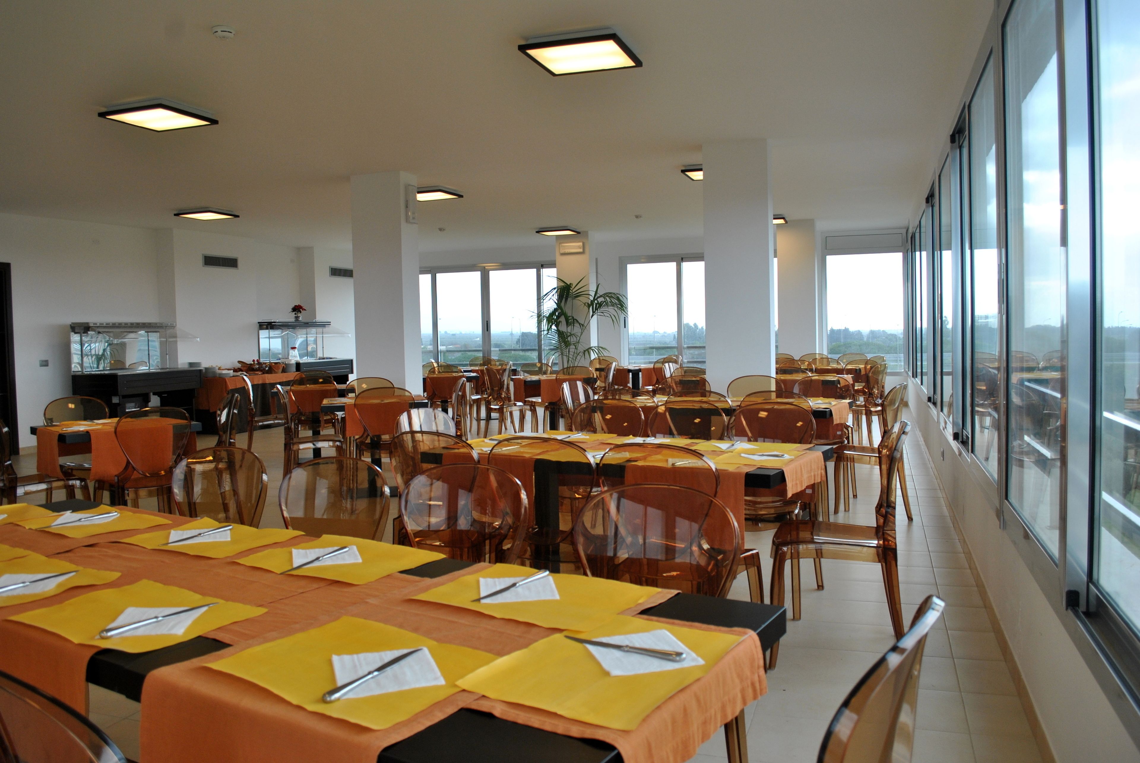 Uma ampla sala de jantar com várias mesas postas, grandes janelas com vista e uma área de cozinha ao fundo.