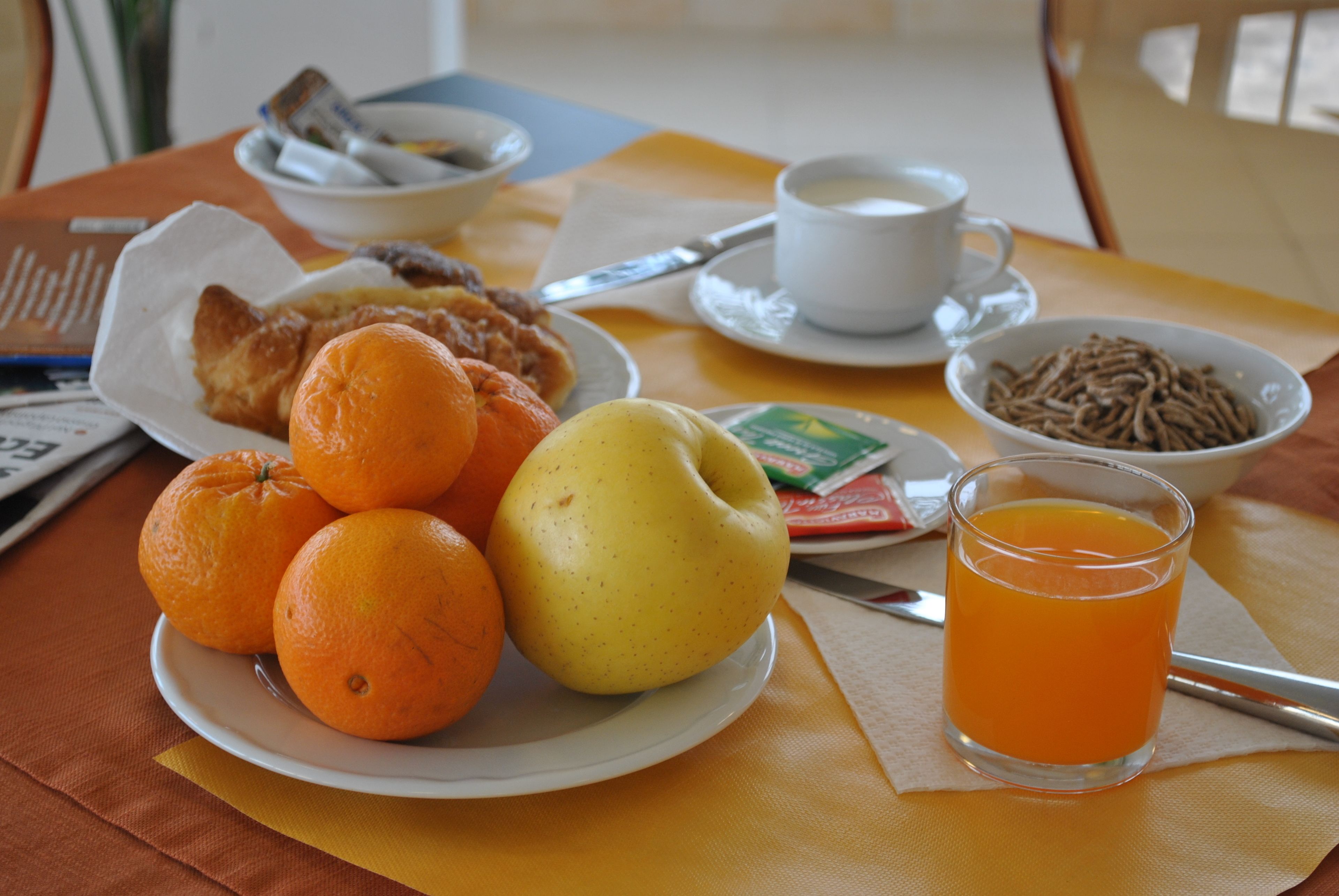 A breakfast table with plates of oranges, a pear, a glass of orange juice, and a bowl of cereal.