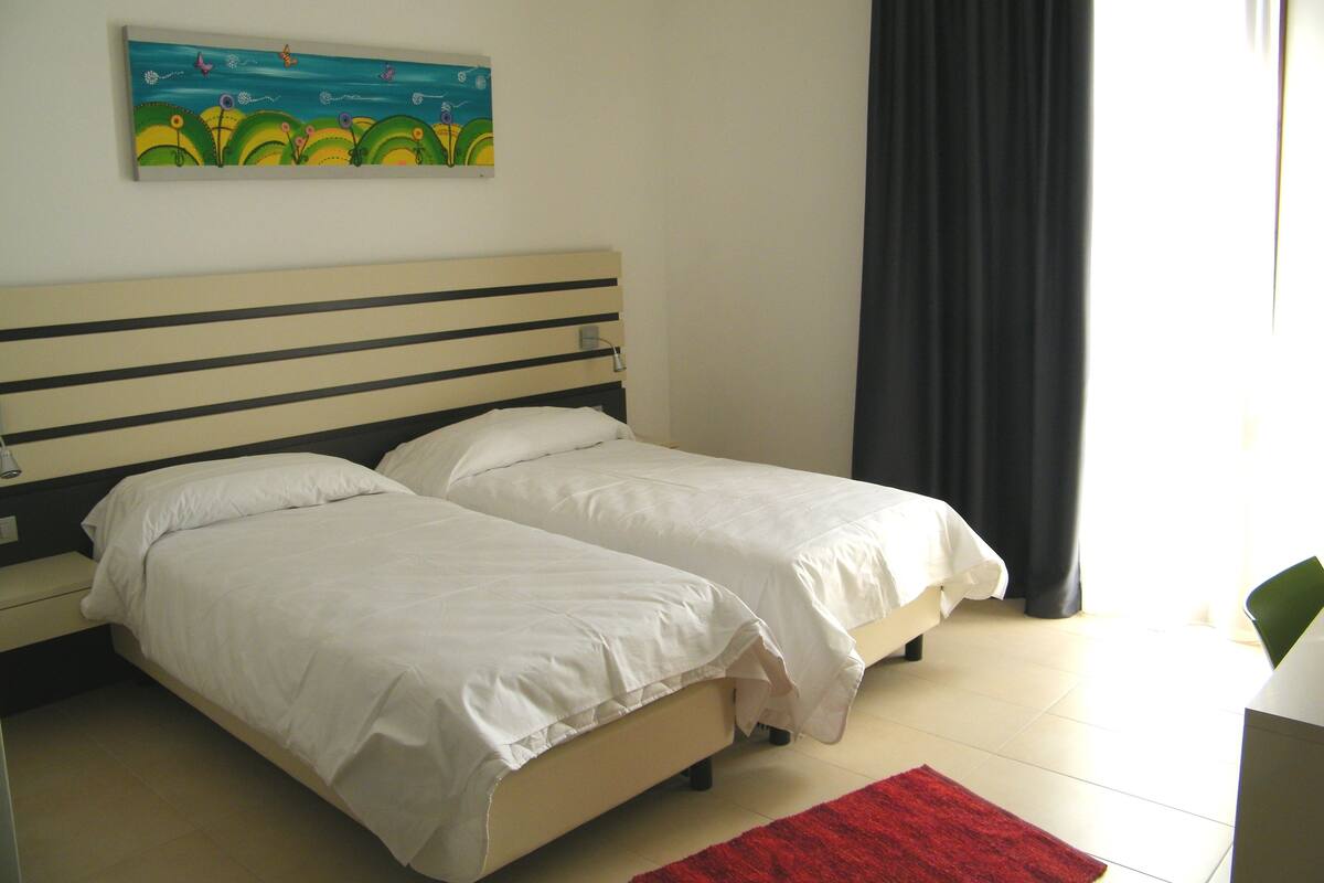 Ein Hotelzimmer mit zwei Einzelbetten, einem Gemälde an der Wand, einem roten Teppich auf dem Boden und einem Fenster mit Vorhängen.
