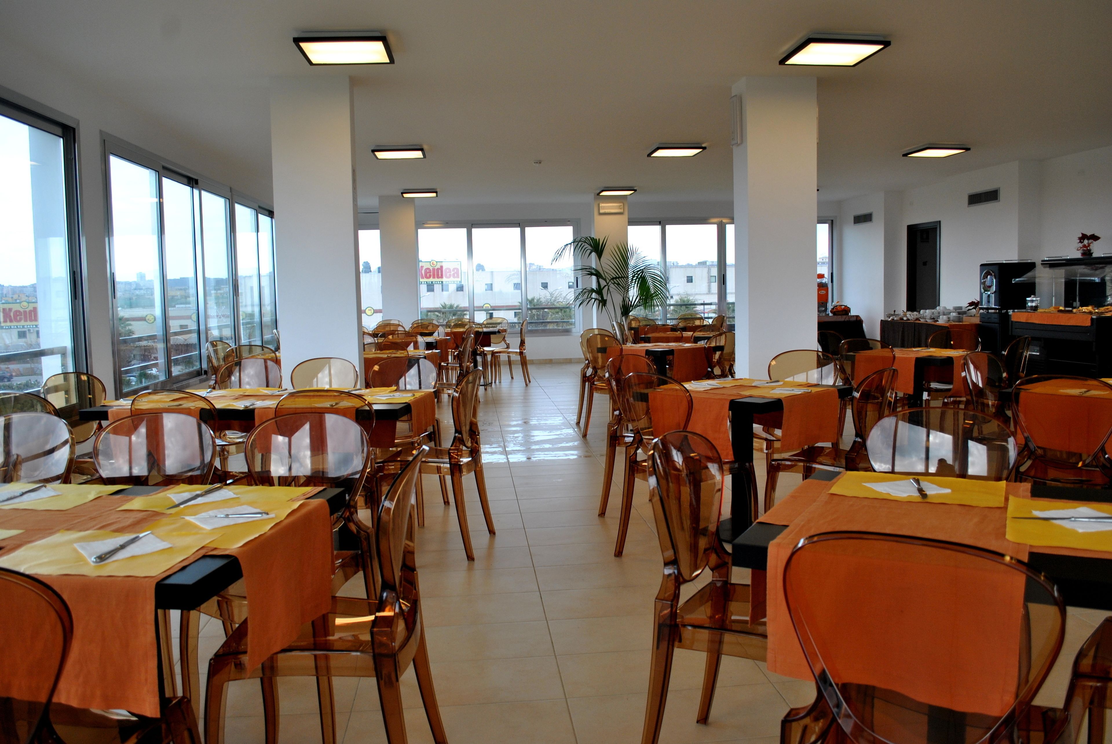 A spacious dining area with multiple tables and chairs, large windows offering views of the city, and a kitchen area visible in the background.