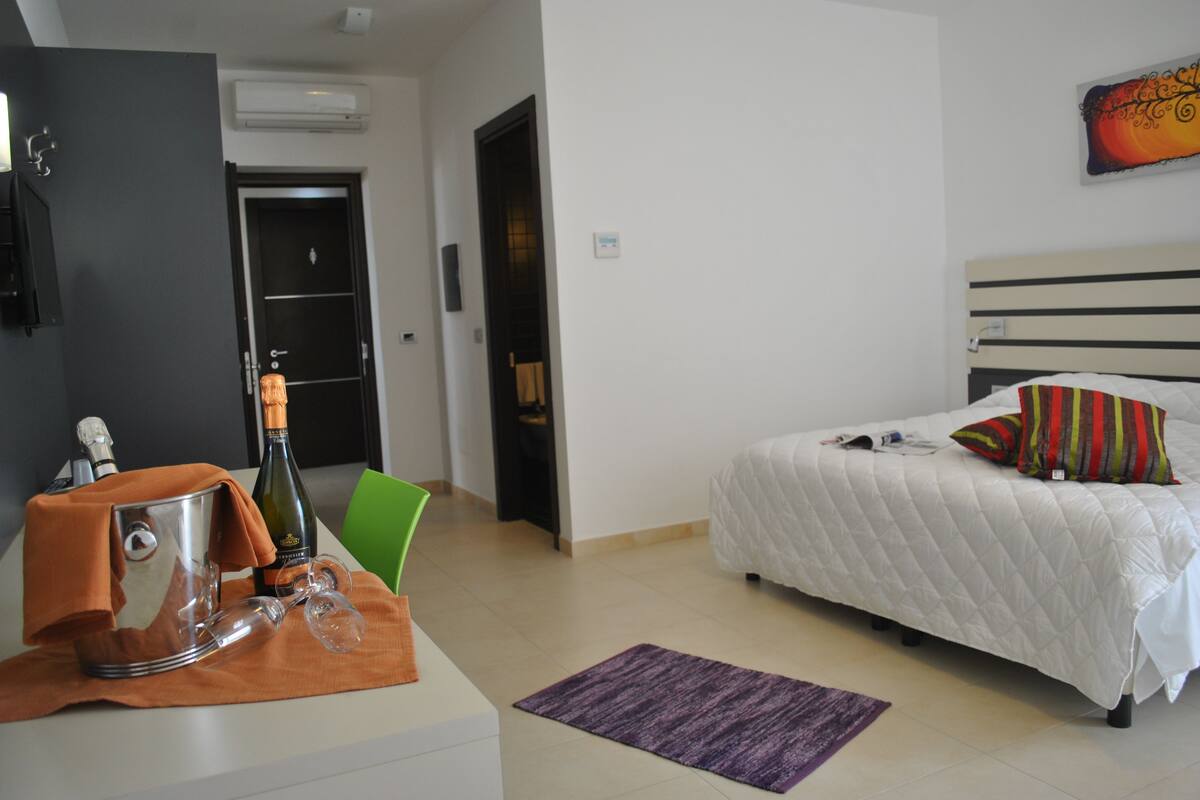 A hotel room with a bed, a nightstand, a chair, a purple rug, and a bottle of wine.