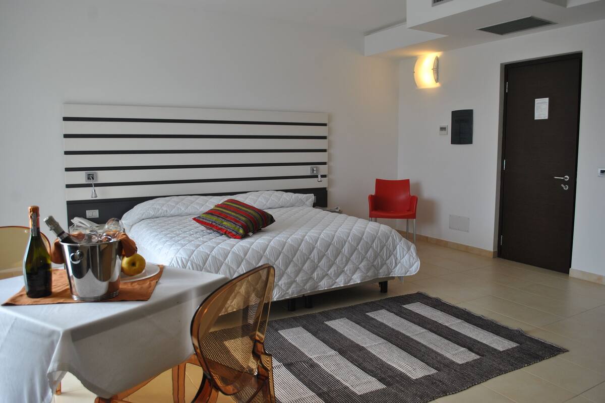 A hotel room with a bed, a table with a bottle and glasses, a red chair, and a door.