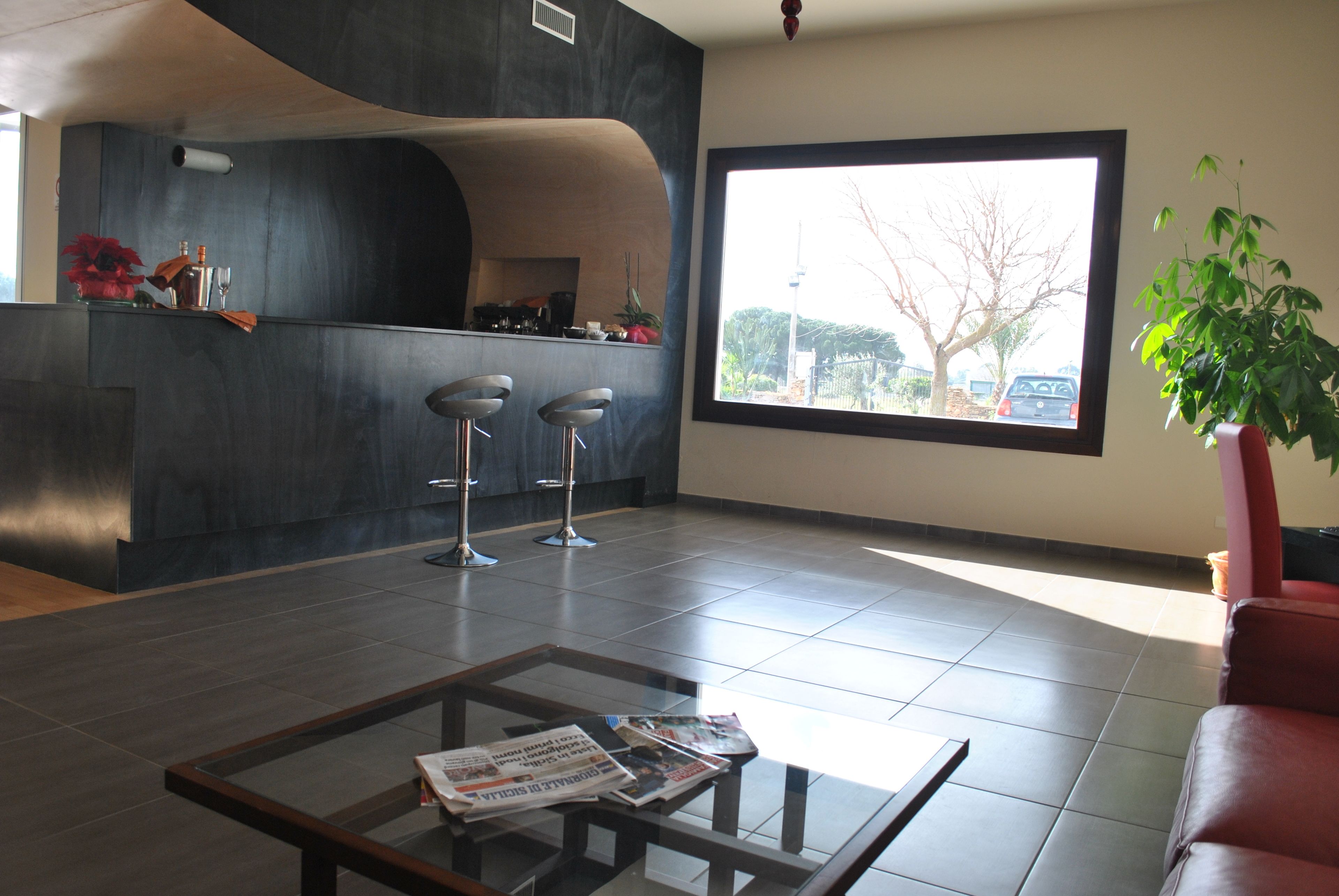 A modern lobby with a glass coffee table, two bar stools, a large window with a view, and a potted plant.
