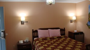 A hotel room with a bed, two bedside lamps, a nightstand with a phone and a lamp, and a wall-mounted heater. - Pinconning Trail Inn Motel (Pinconning)