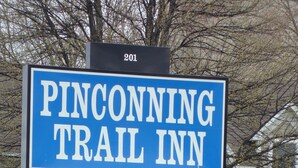 Letrero de motel para el Pinconning Trail Inn, con un letrero azul grande con letras blancas, un letrero negro más pequeño debajo con una bandera estadounidense y un símbolo de WiFi, y una cerca de piquetes blancos al fondo.