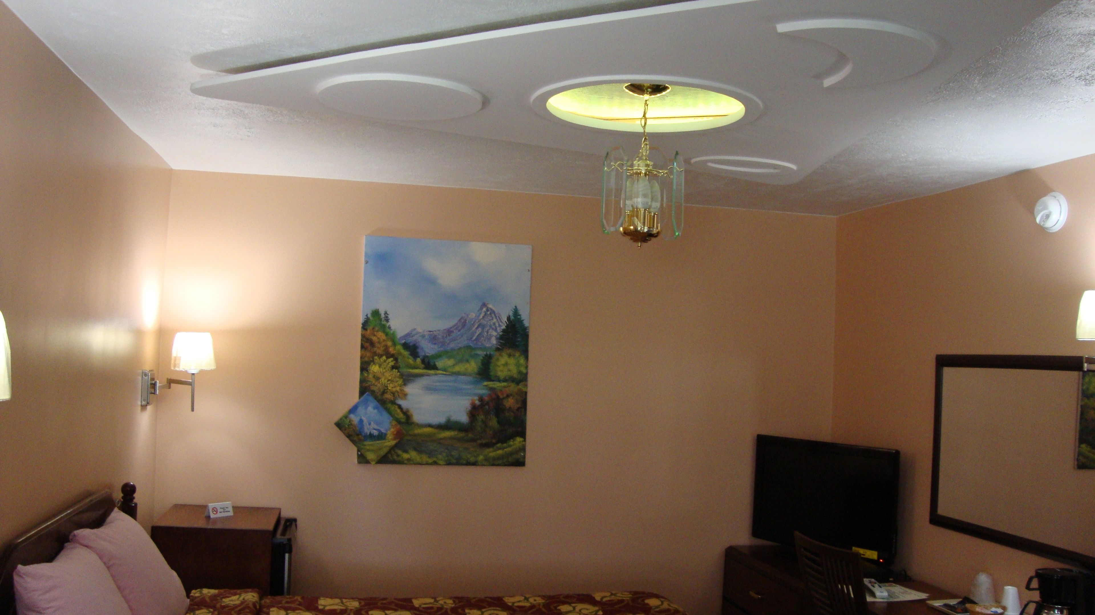 Habitación de hotel con cama, escritorio, silla, un cuadro de un paisaje, una lámpara en el techo y un televisor montado en la pared.