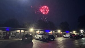 Un estacionamiento con varios vehículos, un edificio con letreros numerados y una exhibición de fuegos artificiales al fondo.