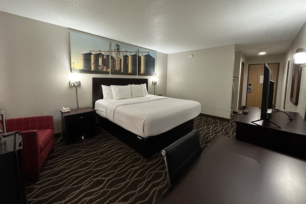 A hotel room with a bed, a desk, a chair, a red sofa, a mirror, and a picture of industrial buildings on the wall.