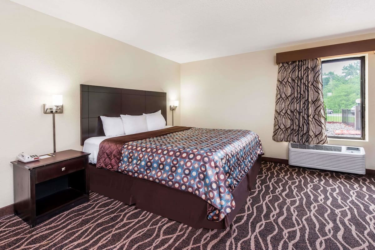 A hotel room with a bed, bedside table, lamp, air conditioning unit, and a window with curtains.