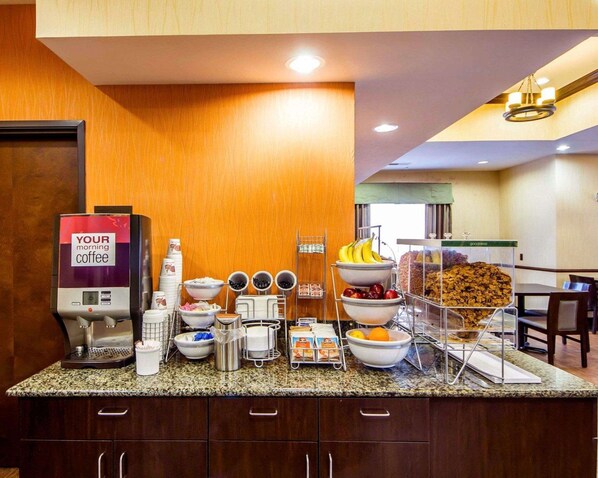 A hotel breakfast area with a coffee machine, bowls of fruits, and a variety of breakfast items on a granite countertop.