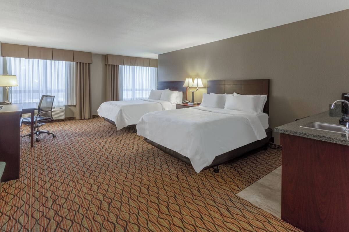 A hotel room with two beds, a desk, a chair, a small desk area, and a bathroom with a sink and mirror.