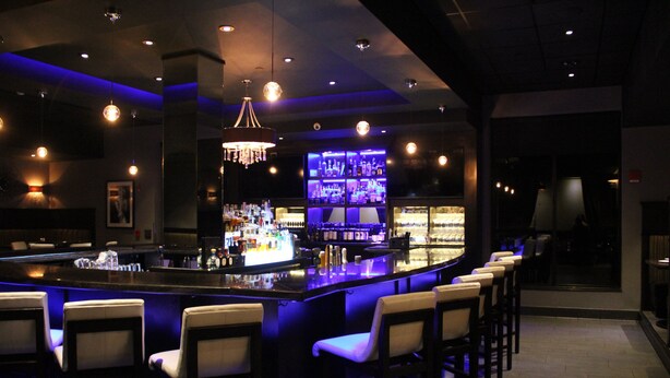 Una zona de bar con una barra alargada, varios taburetes y una vitrina bien iluminada.