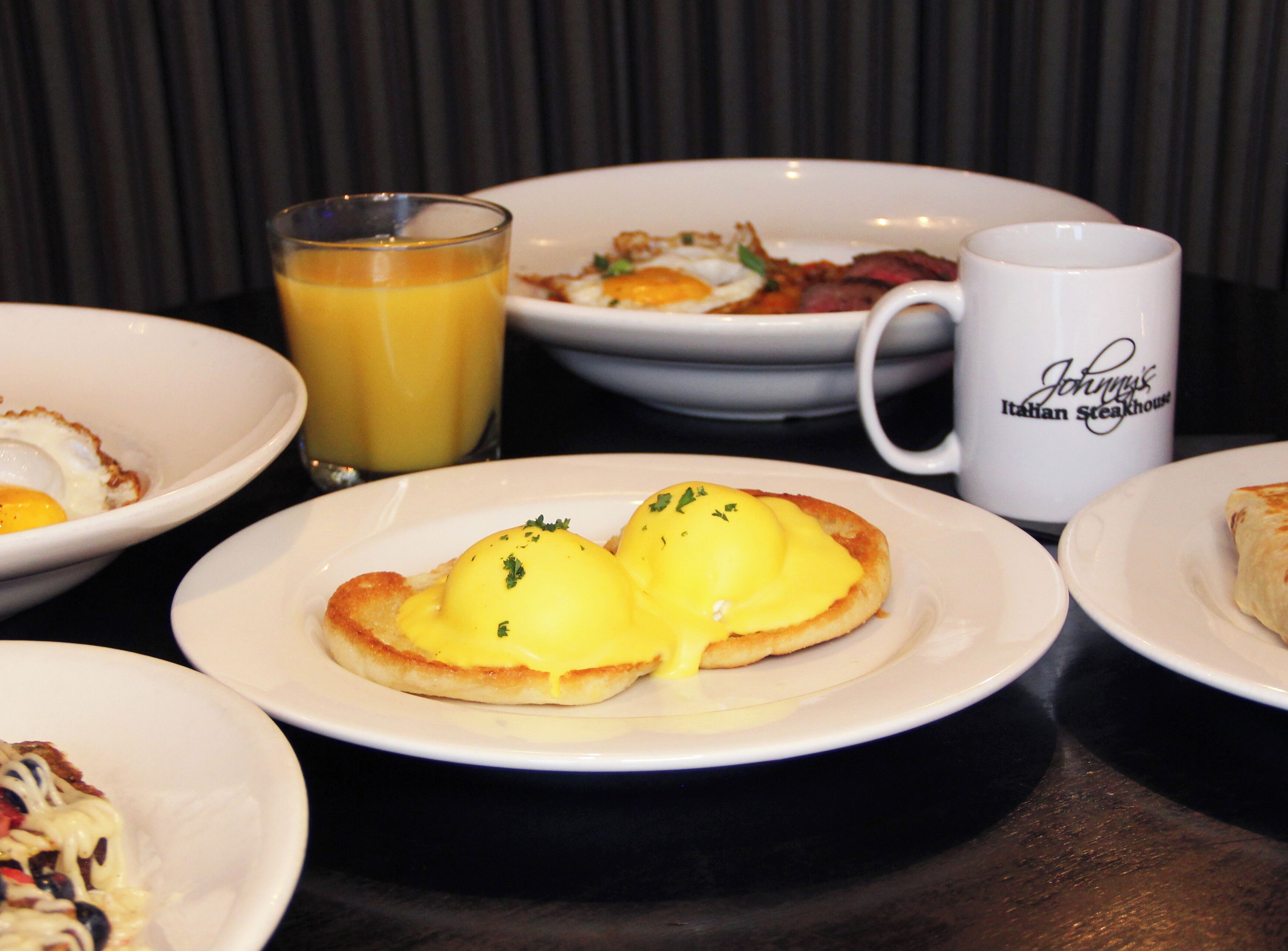 A breakfast setting with various dishes including eggs Benedict, a glass of orange juice, and a mug labeled "Johnny's Italian Steakhouse".