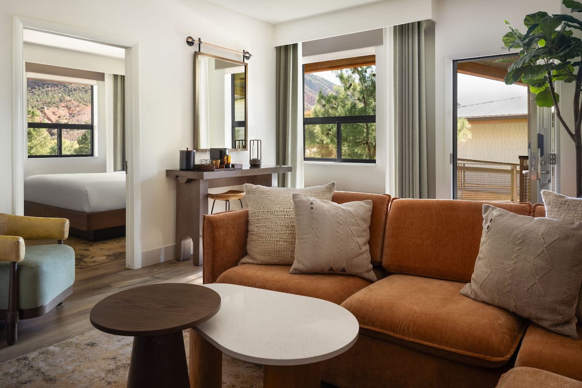A modern hotel room with a sofa, a small table, a desk with a mirror, and large windows offering views of the outdoors.