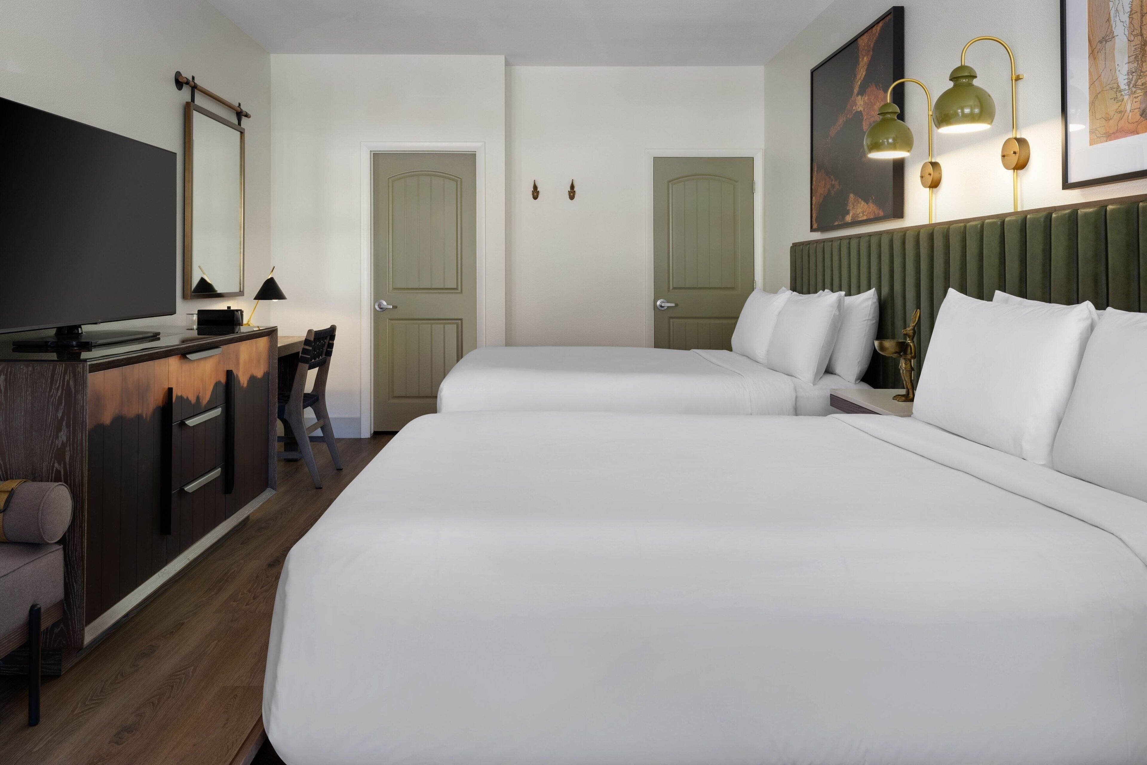 A hotel room with two beds, a desk with a TV, a chair, and a green headboard.
