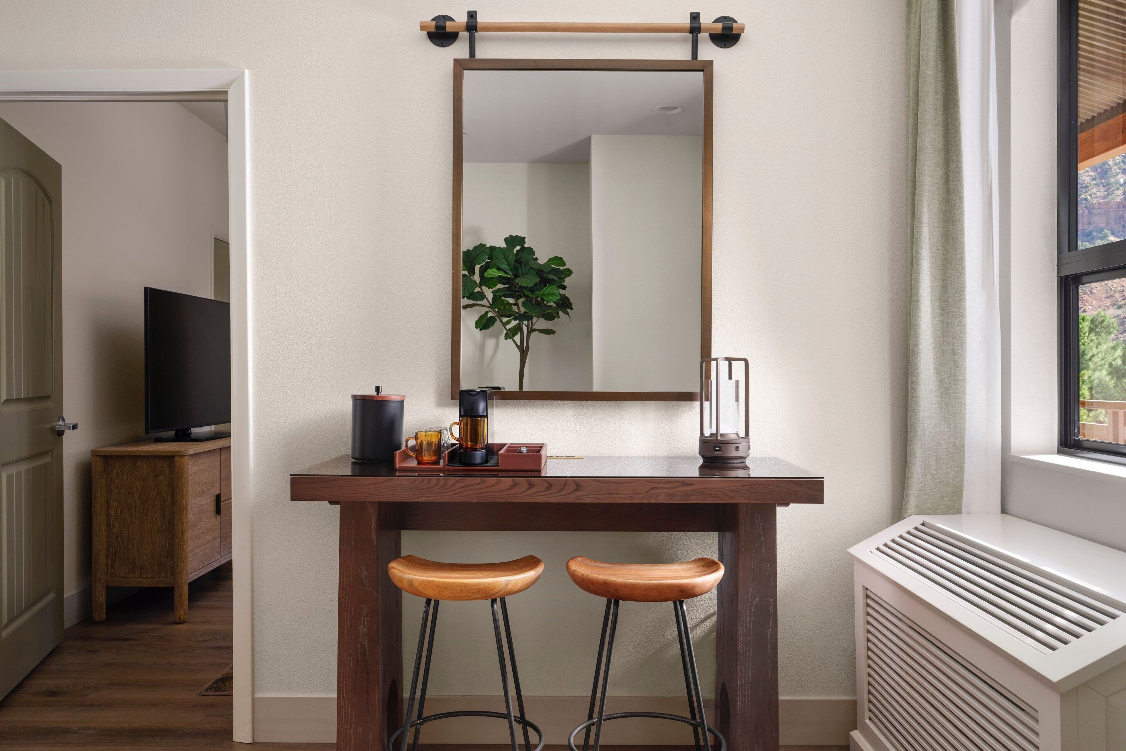 A room with a wooden desk, two stools, a mirror, a plant, a TV, and a radiator.
