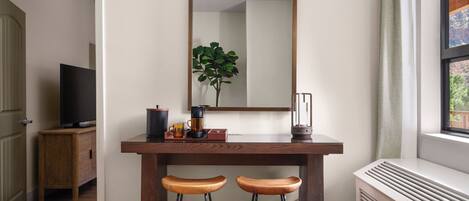 A room with a wooden desk, two stools, a mirror, a plant, a TV, and a radiator.