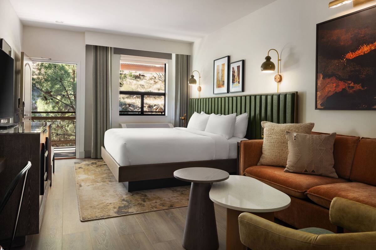 A hotel room with a bed, a sofa, a small table, a TV, and a window with a view of trees.