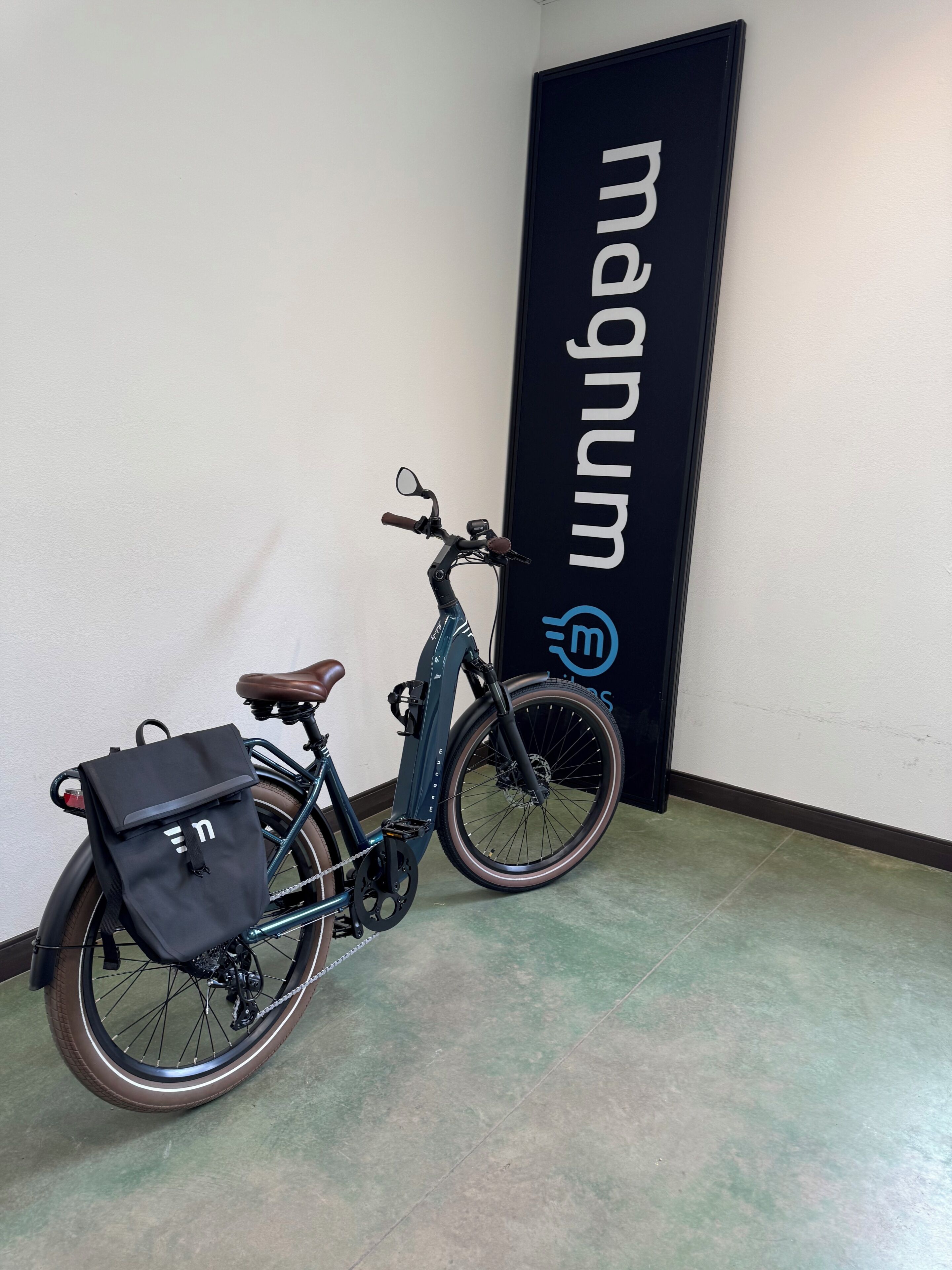 A stationary bicycle with a black bag attached to the rear, positioned against a wall with a vertical sign displaying the word 'Mabtum' and a logo.