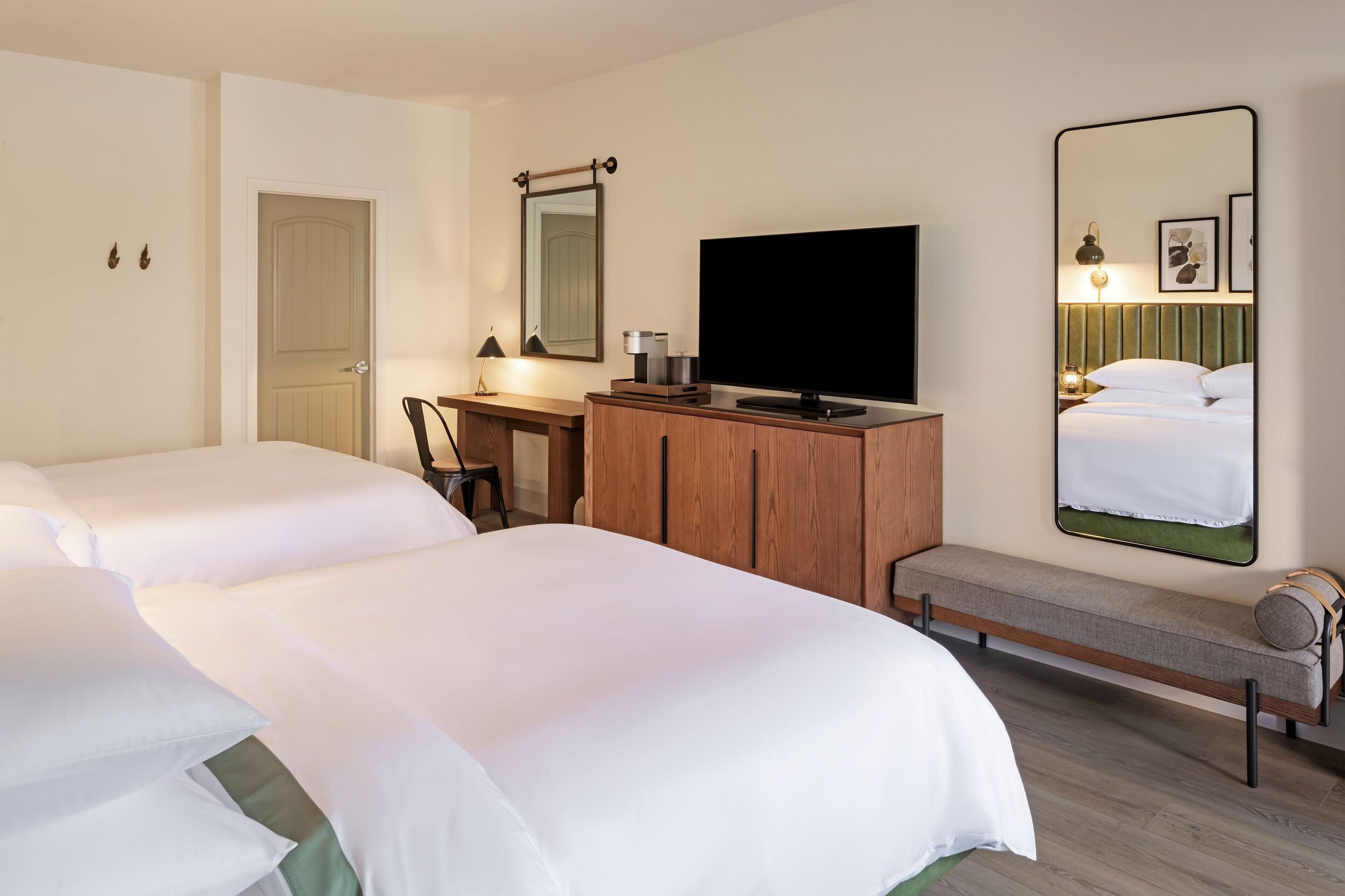 A hotel room with two beds, a TV, a desk with a chair, a mirror, and a bench.
