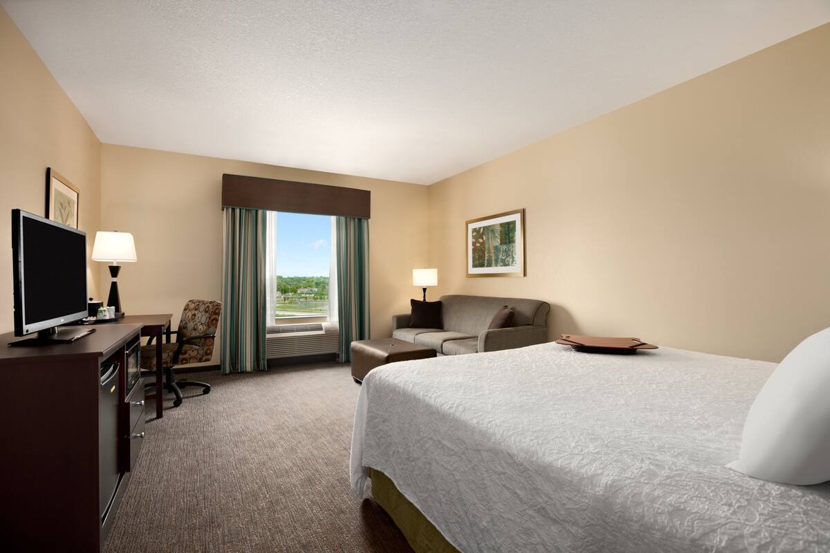 A hotel room with a bed, a desk with a TV, a chair, a sofa, and a window with curtains.
