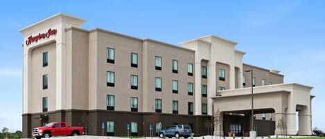 A Hampton Inn hotel with a modern design, featuring a flat roof, large windows, and a parking area with vehicles.