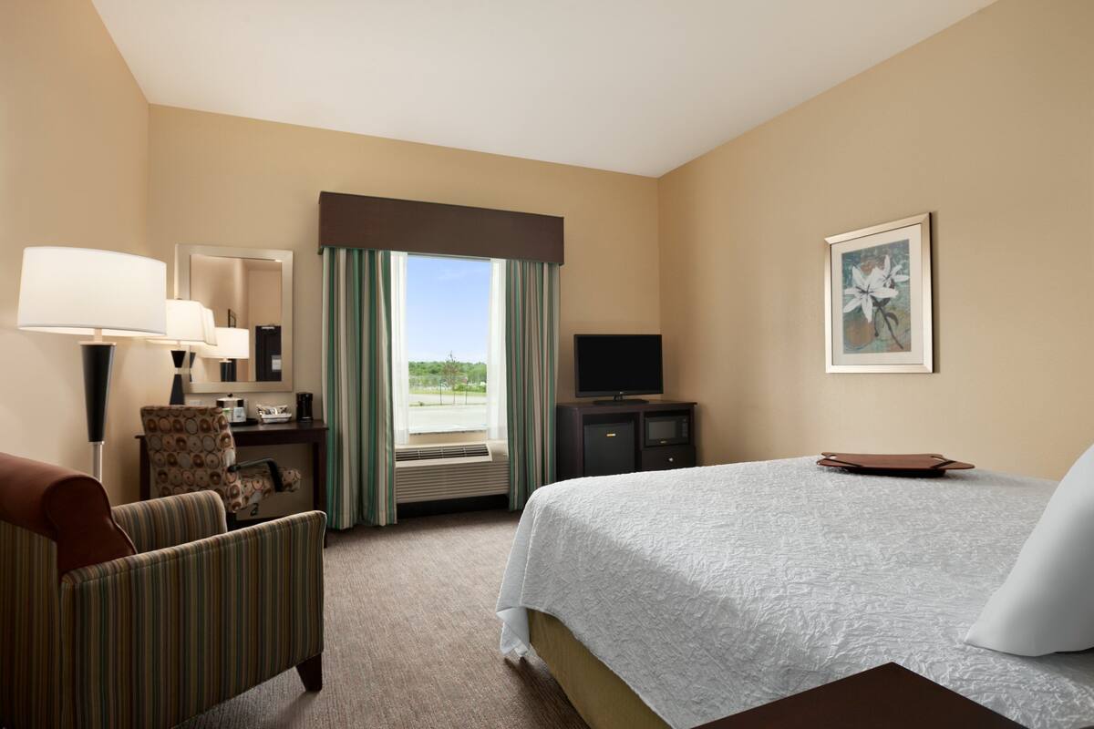 A hotel room with a bed, a desk with a lamp, a chair, a television, and a window with curtains.