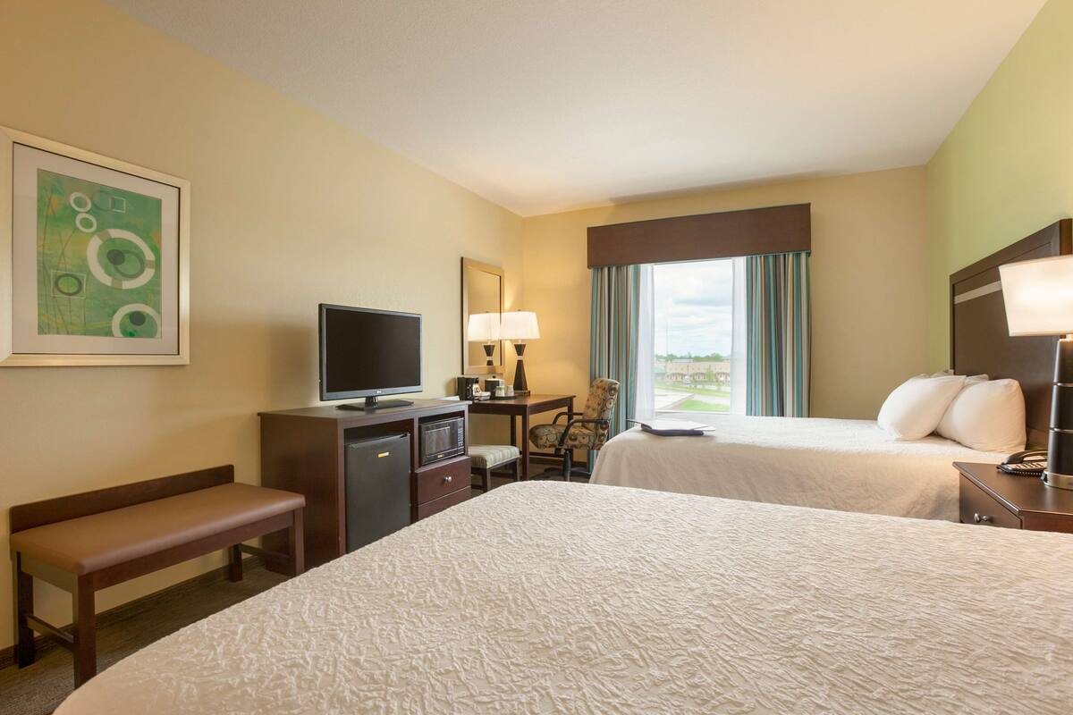 A hotel room with two beds, a desk with a TV, a chair, a bench, and a window with curtains.